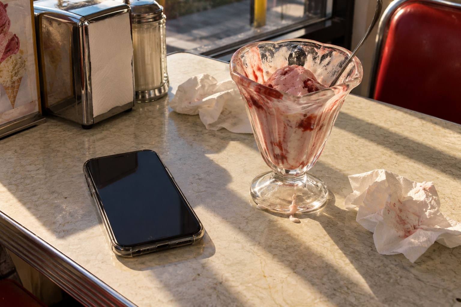 Celular con la pantalla apagada sobre la mesa de una heladería retro, junto a una copa de helado de fresa a medio comer y servilletas arrugadas