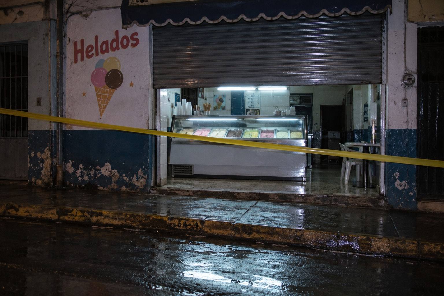 Fachada de una heladería de barrio de noche, con la persiana a medio bajar, la vitrina iluminada y una cinta amarilla cruzando la entrada